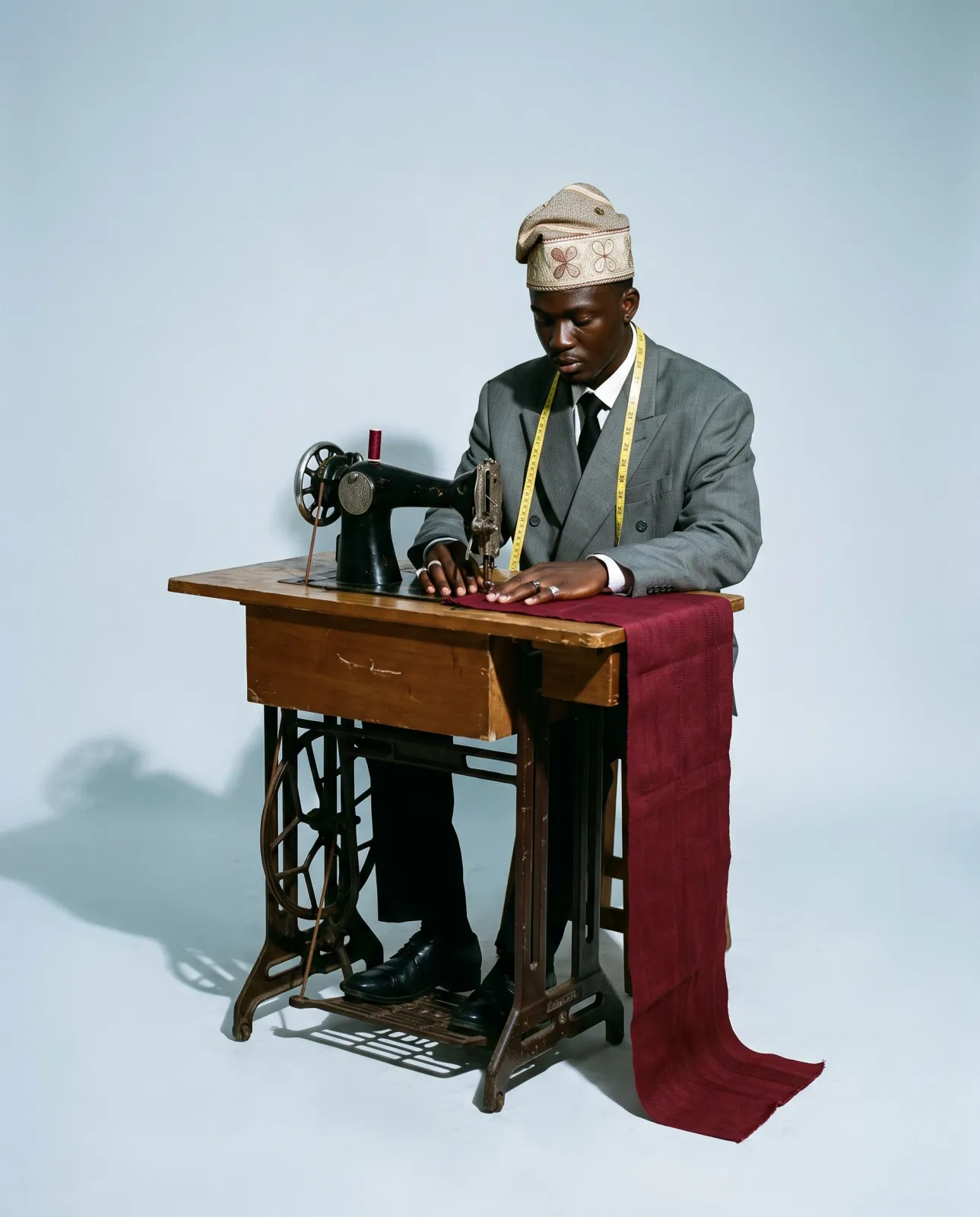 A young Nigerian tailor at an old treadle sewing machine, feeding wine-red aso oke under the needle, a tape measure round his neck
